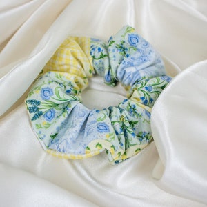 Puede incluir: Una goma para el pelo floral con patrones azules, amarillos y verdes. La goma para el pelo es de tela y tiene un diseño fruncido. El fondo es una tela blanca y sedosa.