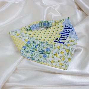 May include: A patchwork pet bandana with a floral and gingham pattern in yellow, blue, and white. The name "Finley" is embroidered in blue on a light blue patch. The bandana is set against a white satin background.