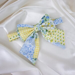 Può includere: Un grande fiocco decorativo in tessuto con un design patchwork. Presenta sezioni di giallo a quadretti, motivi floreali blu e stampe floreali gialle. Il fiocco è posto su uno sfondo bianco e serico.