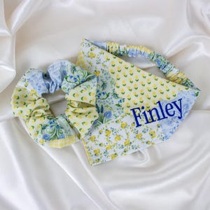 May include: A scrunchie and a headband set with a floral pattern in yellow, blue, and green. The headband is personalized with the name "Finley" in blue lettering. The items are displayed on a white satin surface.