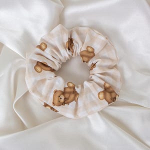 May include: A beige scrunchie with a repeating pattern of brown teddy bears. The fabric has a subtle checkered design. The scrunchie is a hair accessory, perfect for adding a touch of whimsy to any hairstyle.