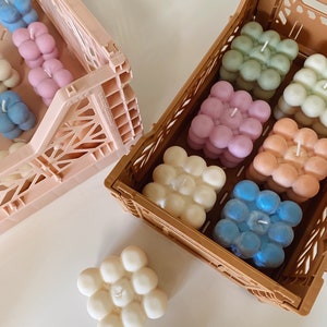 May include: Bubble-shaped candles in various pastel colors, including blue, pink, and green, are arranged in brown and pink plastic crates. One white candle sits outside the crates. The candles have a unique, textured design.