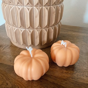 May include: Two small orange pumpkin-shaped candles with white wicks. The candles are sitting on a wooden surface.