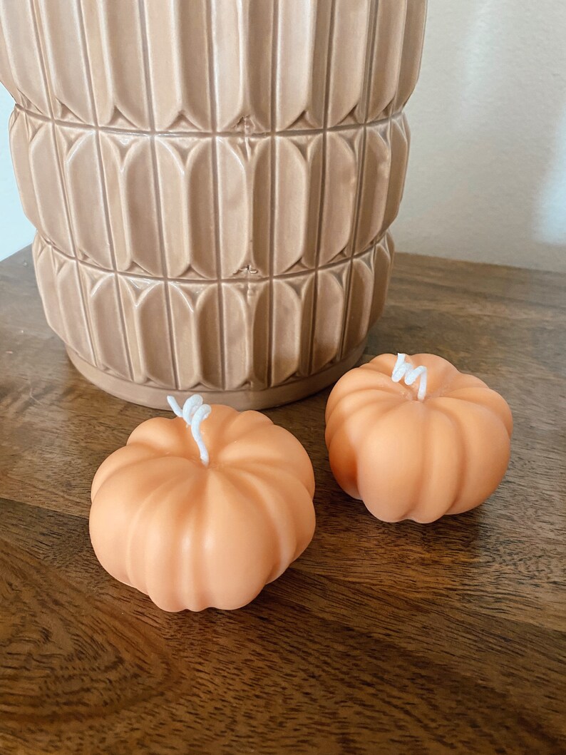 May include: Two small orange pumpkin-shaped candles with white wicks. The candles are sitting on a wooden surface.