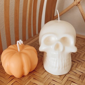 May include: Two Halloween-themed candles, one shaped like a pumpkin and the other shaped like a skull. The pumpkin candle is orange and the skull candle is white. Both candles are sitting on a woven bamboo surface.