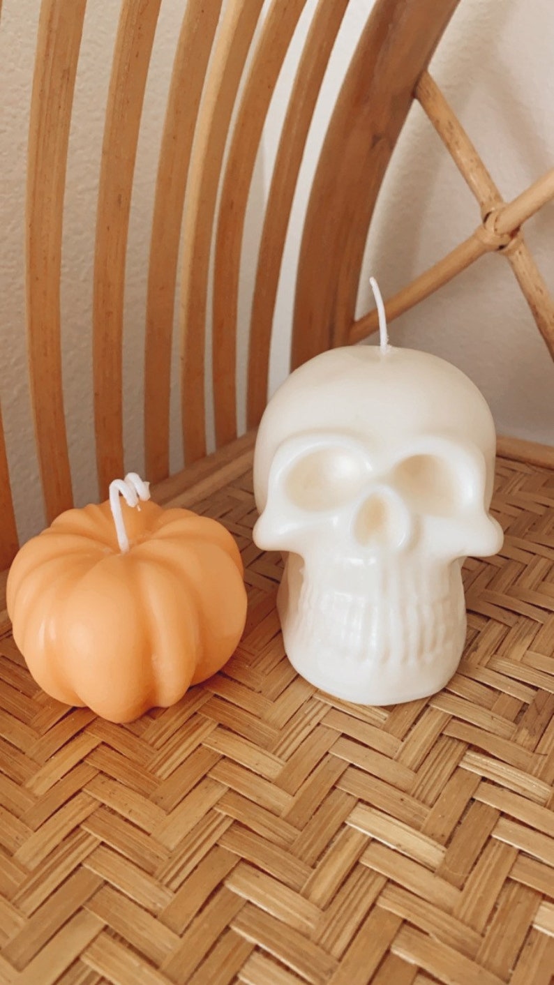 May include: Two Halloween-themed candles, one shaped like a pumpkin and the other shaped like a skull. The pumpkin candle is orange and the skull candle is white. Both candles are sitting on a woven bamboo surface.
