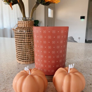 May include: Two orange pumpkin-shaped candles and a red candle with a white plus sign pattern. The red candle is in a glass jar.