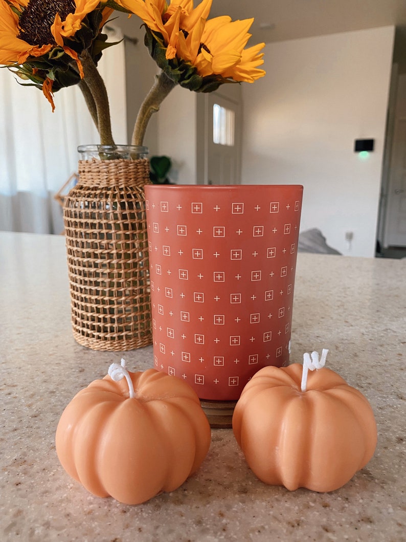 May include: Two orange pumpkin-shaped candles and a red candle with a white plus sign pattern. The red candle is in a glass jar.