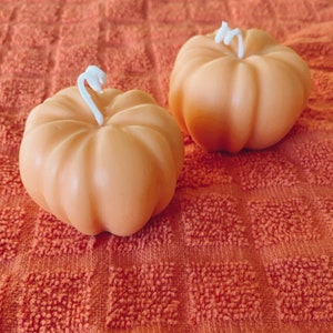 May include: Two small orange pumpkin-shaped candles with white wicks on a textured orange surface.