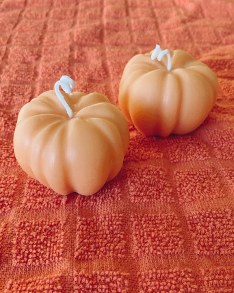 May include: Two small orange pumpkin-shaped candles with white wicks on a textured orange surface.
