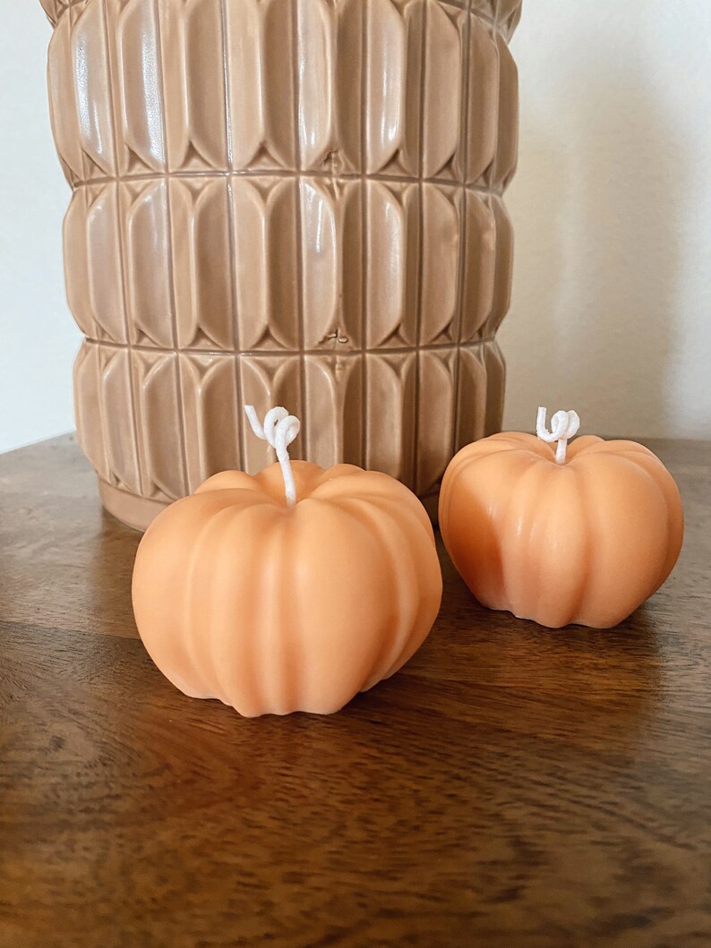 May include: Two small orange pumpkin-shaped candles on a wooden surface. The candles have white wicks.