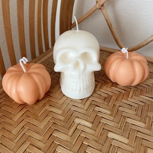 May include: Three Halloween-themed candles: a white skull candle and two orange pumpkin candles. The candles are arranged on a woven wicker surface.