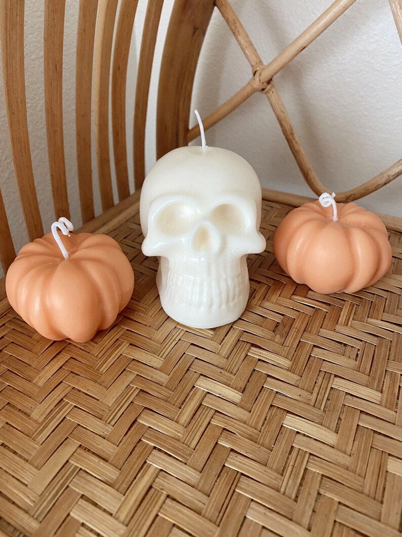 May include: Three Halloween-themed candles: a white skull candle and two orange pumpkin candles. The candles are arranged on a woven wicker surface.