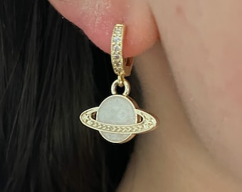 Puede incluir: Aretes de aro dorados con un dije de planeta blanco. El aro está incrustado con pequeñas piedras transparentes.