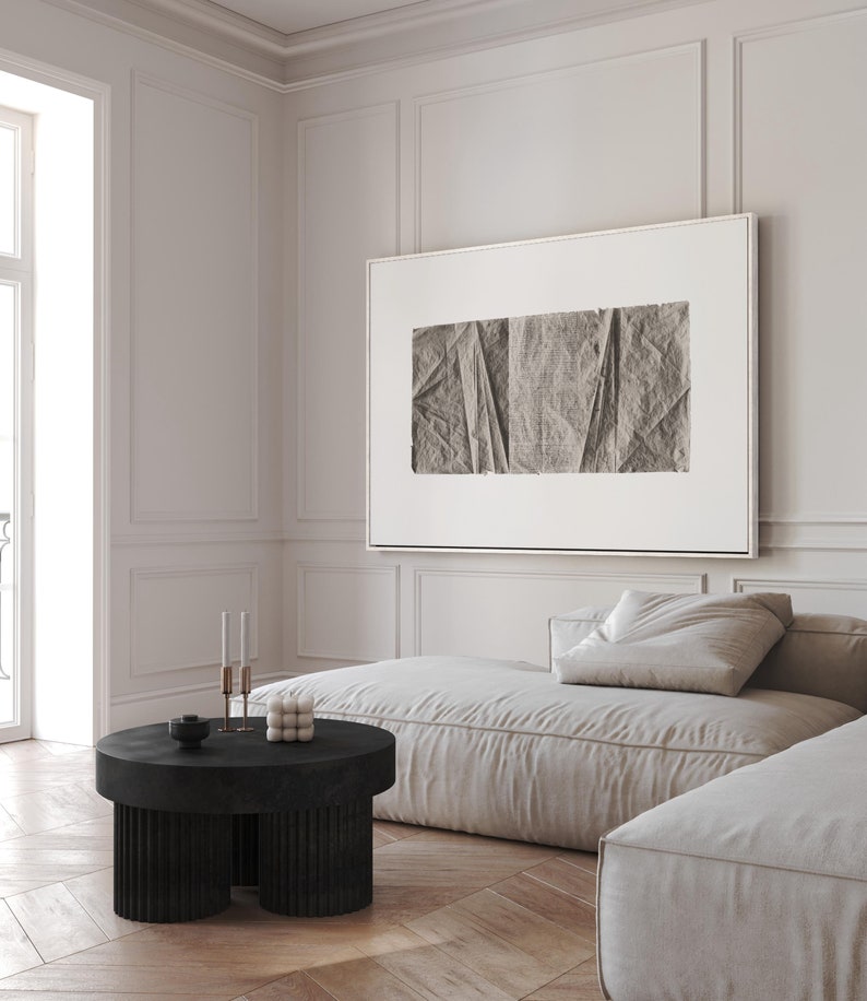 K&ouml;nnte beinhalten: Ein wei&szlig;es, geteiltes Sofa mit einem gro&szlig;en, gerahmten abstrakten Druck dar&uuml;ber. Das Sofa steht in einem Raum mit wei&szlig;en W&auml;nden und einem Holzboden. Ein schwarzer, runder Couchtisch mit einem geriffelten Untergestell steht vor dem Sofa.