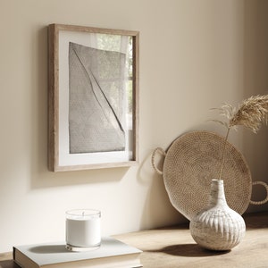 May include: A framed black and white abstract print with a woven tray and a white ceramic vase with a dried plant on a wooden surface.