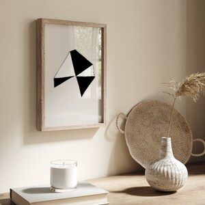 May include: A framed black and white geometric abstract art print with a wooden frame. The print is hanging on a beige wall next to a wicker tray and a white ceramic vase with a dried flower arrangement.