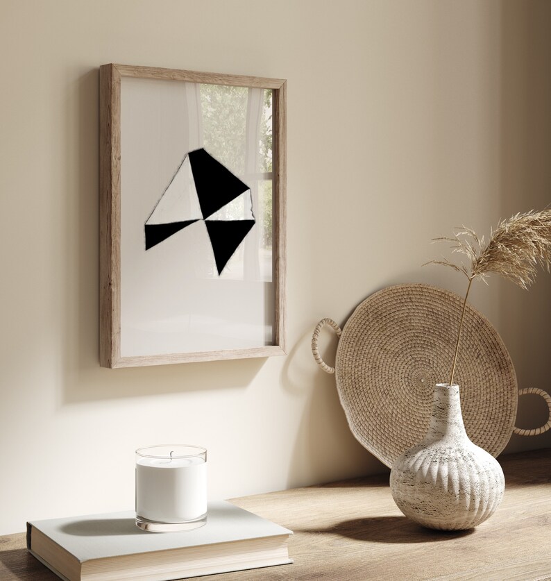 May include: A framed black and white geometric abstract art print with a wooden frame. The print is hanging on a beige wall next to a wicker tray and a white ceramic vase with a dried flower arrangement.