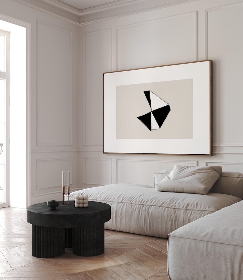 May include: A black and white abstract geometric art print in a wooden frame hangs above a light beige sectional sofa with a black round coffee table. The coffee table has three legs and a textured surface.