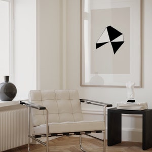 May include: A white leather armchair with a black metal frame sits in a room with white walls and hardwood floors. A black and white abstract print hangs on the wall above a black side table with books and a white sculpture.
