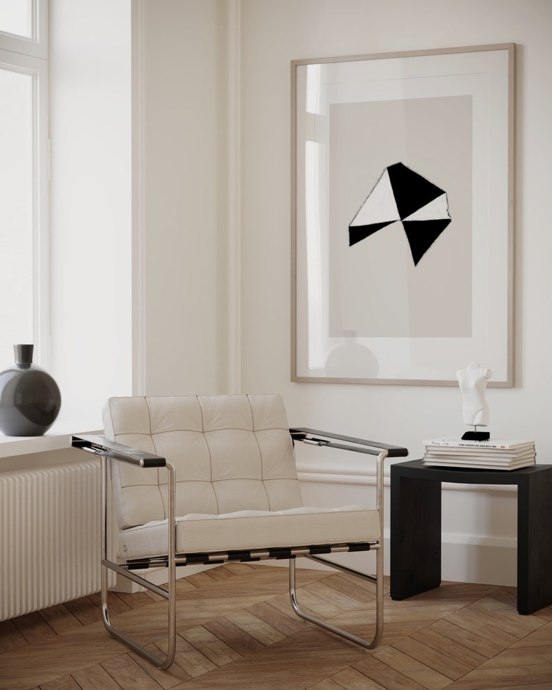 May include: A white leather armchair with a black metal frame sits in a room with white walls and hardwood floors. A black and white abstract print hangs on the wall above a black side table with books and a white sculpture.