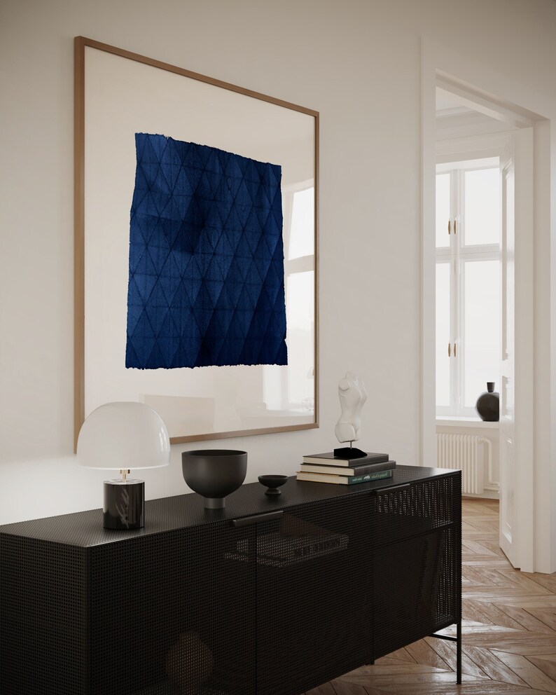 May include: A black cabinet with a wire mesh front and a white lamp with a white shade on top. There is a blue framed artwork with a blue geometric pattern on the wall behind the cabinet.