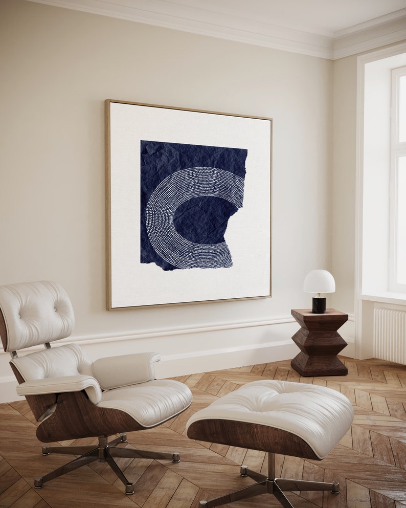 May include: A white leather armchair and ottoman with a dark wood base. A framed abstract art print with a navy blue background and white dots hangs on the wall behind the chair.