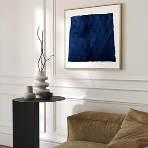 May include: A blue abstract painting with a geometric pattern in a gold frame hanging on a white wall above a black side table with a vase and two decorative objects. A brown sofa is in the foreground.