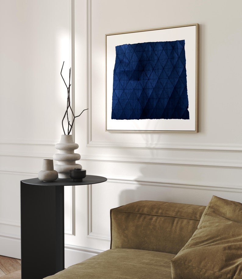May include: A blue abstract painting with a geometric pattern in a gold frame hanging on a white wall above a black side table with a vase and two decorative objects. A brown sofa is in the foreground.