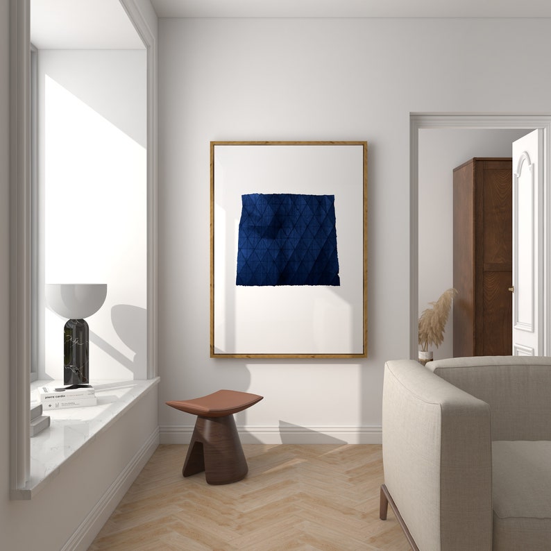 May include: A framed abstract art print with a blue geometric design on a white background. The print is hanging on a white wall in a living room setting with a wooden stool and a white sofa.