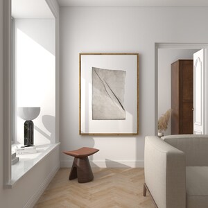 May include: A white living room with a beige sofa, a wooden stool, and a framed print of a folded piece of fabric. The room has a large window and a doorway leading to another room.