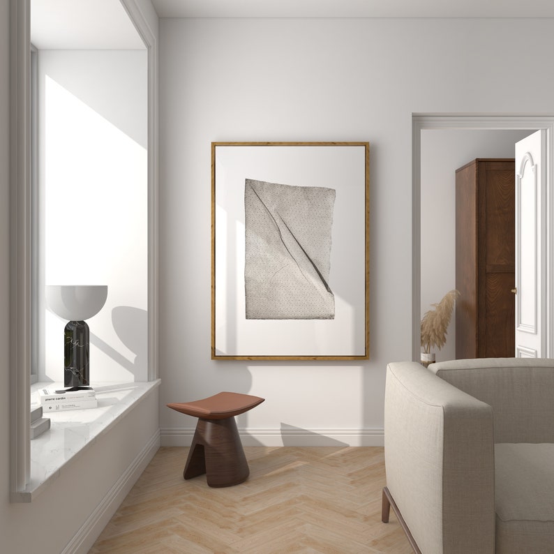 May include: A white living room with a beige sofa, a wooden stool, and a framed print of a folded piece of fabric. The room has a large window and a doorway leading to another room.