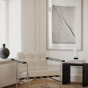 May include: A white leather armchair with a chrome frame sits in a room with white walls and wood floors. A framed print with a black and white abstract design hangs on the wall. A small black side table with a stack of books and a white sculpture sits next to the chair.