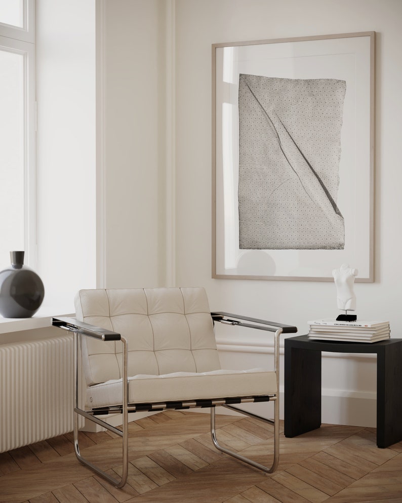 May include: A white leather armchair with a chrome frame sits in a room with white walls and wood floors. A framed print with a black and white abstract design hangs on the wall. A small black side table with a stack of books and a white sculpture sits next to the chair.