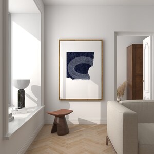 May include: A minimalist living room interior with a white wall, a wooden floor, a large window, a brown leather stool, a white couch, and a framed print of a blue abstract design.