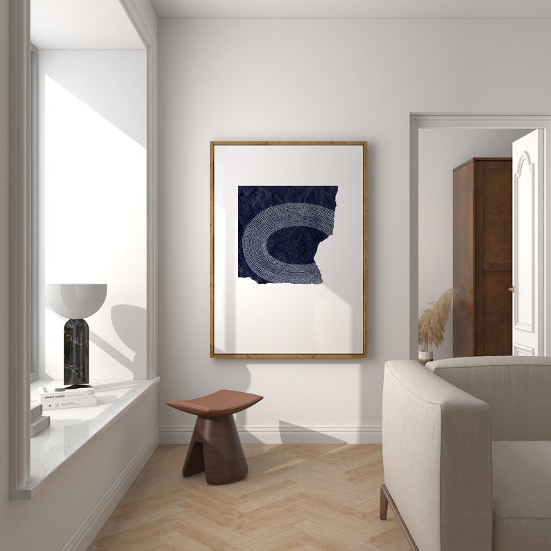 May include: A minimalist living room interior with a white wall, a wooden floor, a large window, a brown leather stool, a white couch, and a framed print of a blue abstract design.