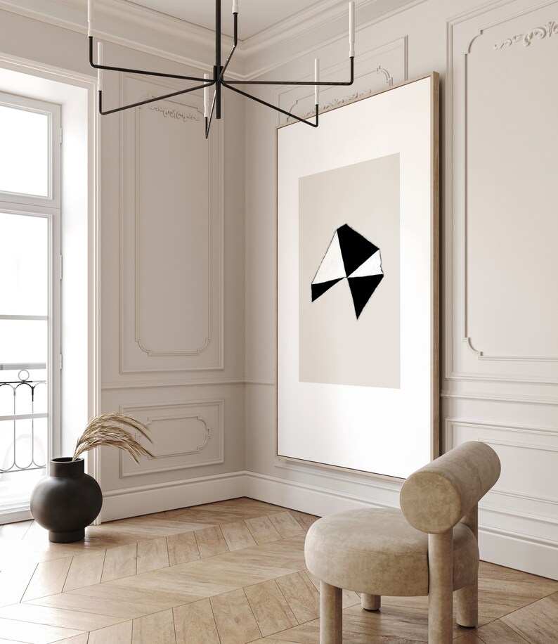 May include: A black and white abstract geometric art print in a simple wooden frame hangs on a white wall in a modern living room. A beige armchair sits in the corner of the room.