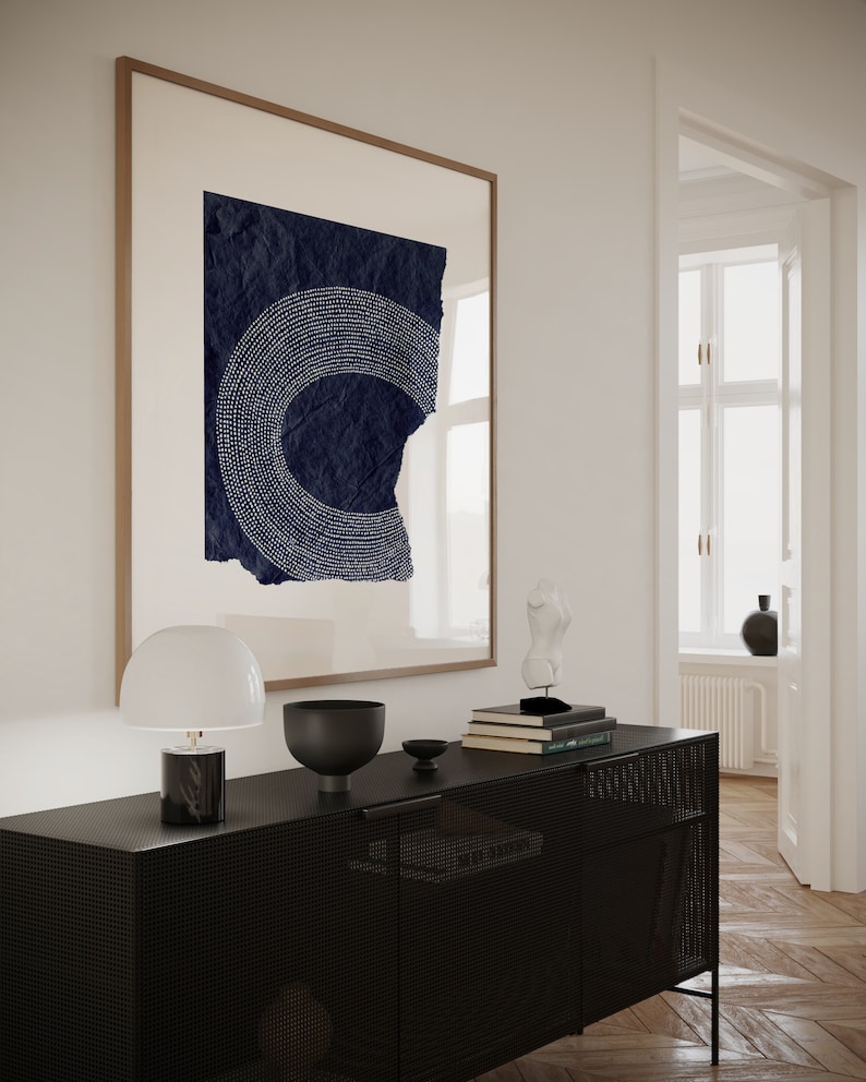 May include: A black metal cabinet with a wire mesh front and a white lamp on the left side. The cabinet is decorated with a black bowl, a stack of books, and a white sculpture. A framed print with a blue and white abstract design hangs on the wall behind the cabinet.
