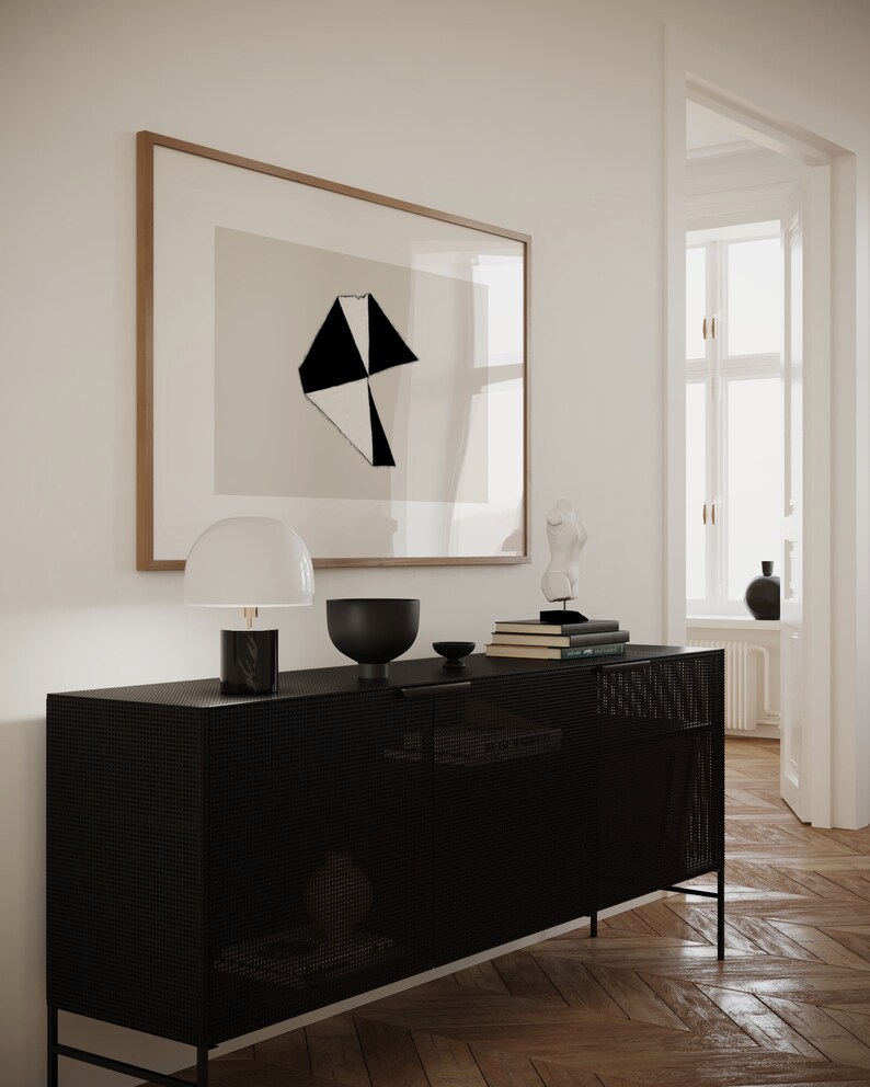 May include: A black metal cabinet with a wire mesh front and two doors. The cabinet is sitting on a wooden floor and has a lamp, a bowl, and books on top of it. There is a framed print of a black and white geometric design on the wall behind the cabinet.
