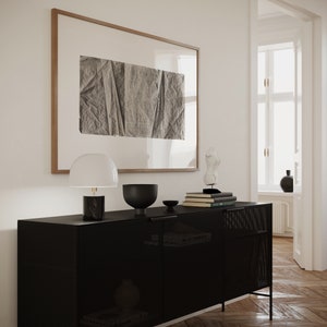 K&ouml;nnte beinhalten: Ein schwarzer Metallschrank mit einer Mesh-Front und zwei T&uuml;ren. Der Schrank steht auf einem Holzboden und hat eine wei&szlig;e Lampe, eine schwarze Sch&uuml;ssel und einen Stapel B&uuml;cher darauf. An der Wand hinter dem Schrank h&auml;ngt ein gerahmtes Schwarzwei&szlig;bild.