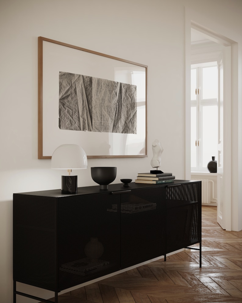 K&ouml;nnte beinhalten: Ein schwarzer Metallschrank mit einer Mesh-Front und zwei T&uuml;ren. Der Schrank steht auf einem Holzboden und hat eine wei&szlig;e Lampe, eine schwarze Sch&uuml;ssel und einen Stapel B&uuml;cher darauf. An der Wand hinter dem Schrank h&auml;ngt ein gerahmtes Schwarzwei&szlig;bild.