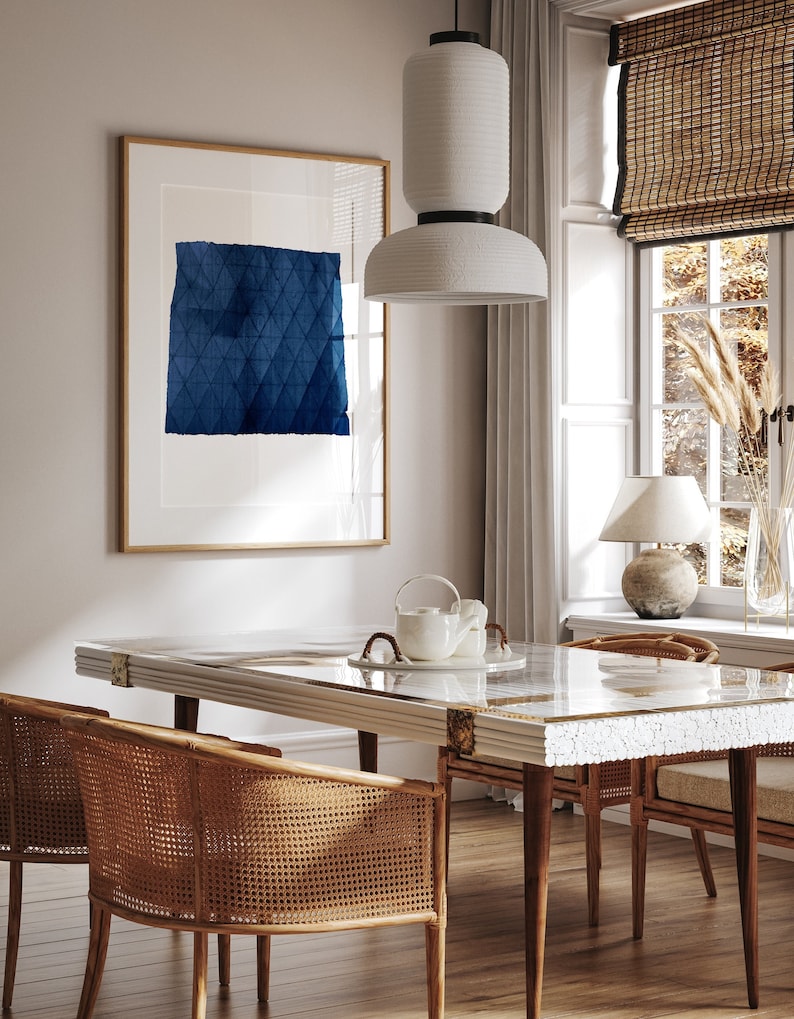 May include: A dining room table with a white marble top and wooden legs. The table is set with a white teapot and two wicker chairs. A large framed print with a blue geometric design hangs on the wall behind the table.