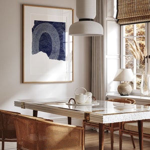 May include: A dining room with a white table with a white and gold inlay, four brown wicker chairs, a white ceramic pendant light, and a framed print with a blue and white abstract design.