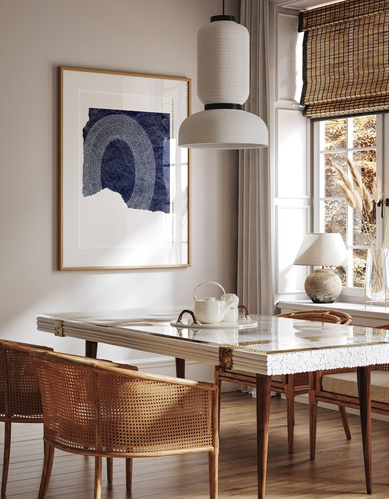 May include: A dining room with a white table with a white and gold inlay, four brown wicker chairs, a white ceramic pendant light, and a framed print with a blue and white abstract design.