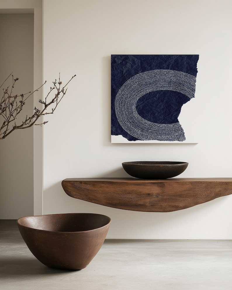 May include: Two brown wooden bowls, one on a wooden shelf and one on the floor, in front of a white wall with a blue and white abstract art print hanging on it.