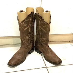 May include: A pair of brown leather cowboy boots featuring a square toe, a stacked heel, and decorative light-coloured stitching patterns on the shaft. Each boot includes a beige plastic boot shaper insert to maintain the upright structure.