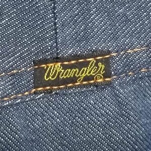 Peut inclure: Gros plan sur un tissu denim bleu fonc&eacute; avec une bordure marron cousue. Une &eacute;tiquette noire rectangulaire avec le mot "Wrangler" en script jaune est cousue sur le denim.