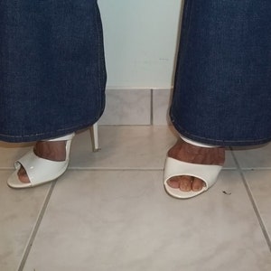 Peut inclure: Sandales blanches &agrave; talons hauts &agrave; bout ouvert. Les sandales ont une seule bride sur le dessus du pied et un talon fin. Les sandales sont port&eacute;es avec un jean en denim bleu.