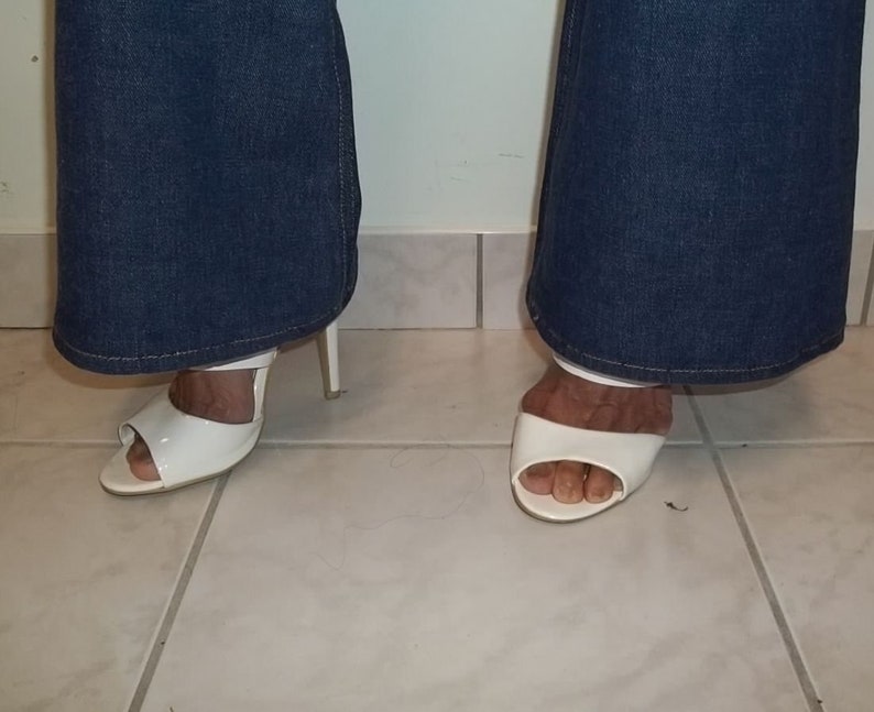 Peut inclure: Sandales blanches &agrave; talons hauts &agrave; bout ouvert. Les sandales ont une seule bride sur le dessus du pied et un talon fin. Les sandales sont port&eacute;es avec un jean en denim bleu.