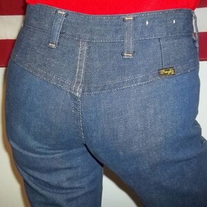 Peut inclure: Gros plan sur un jean en denim bleu avec une taille haute et une &eacute;tiquette Wrangler sur la poche arri&egrave;re. Le jean a une conception classique &agrave; cinq poches avec des coutures blanches et des passants de ceinture. Le tissu semble &ecirc;tre un denim de d&eacute;lavage moyen.
